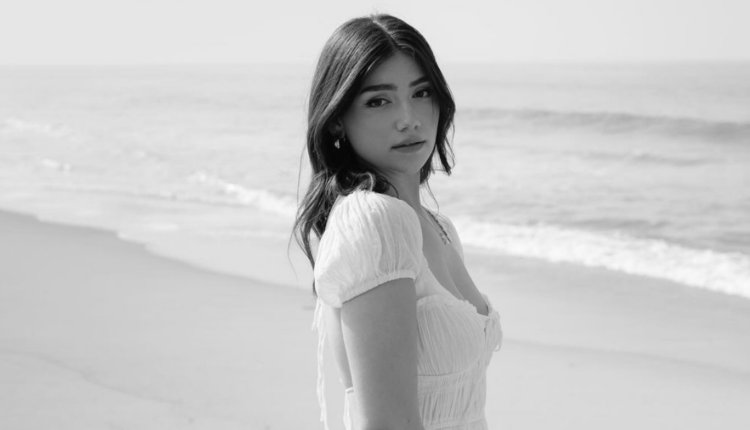 A black and white photo of a young white woman standing on the beach, wearing a white dress and posing in a side profile.